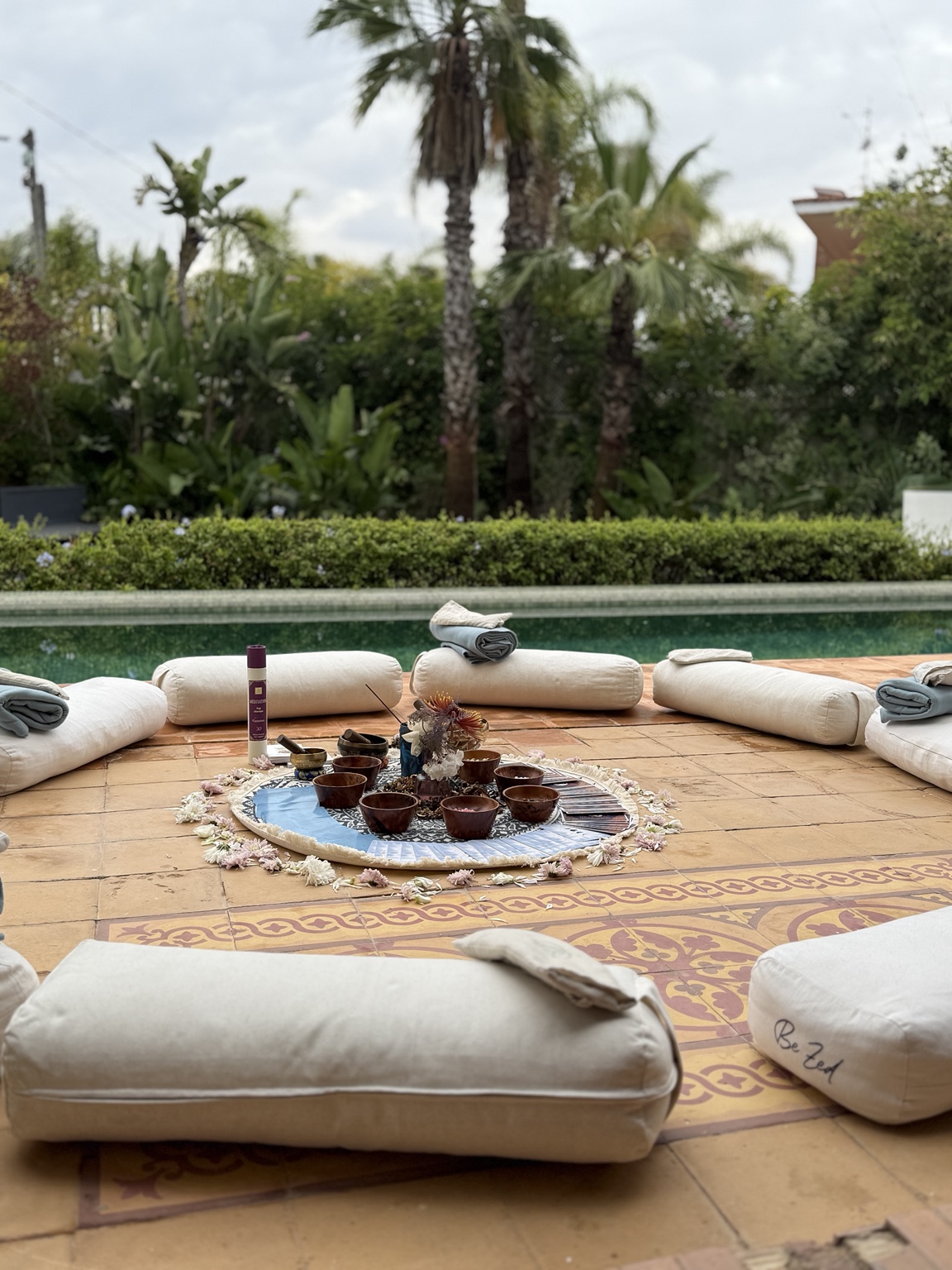Un cercle de coussins autour de fleurs et de petits objets de rituel, au bord d’une piscine